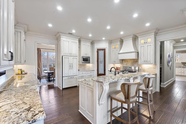 kitchen featuring light stone countertops, premium range hood, built in appliances, a center island with sink, and white cabinets