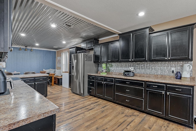 kitchen featuring stainless steel refrigerator with ice dispenser, light countertops, light wood-style flooring, decorative backsplash, and dark cabinets