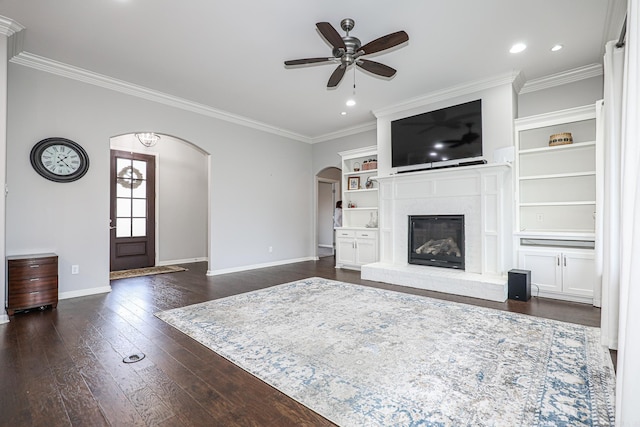 unfurnished living room with a brick fireplace, built in shelves, ceiling fan, dark wood-type flooring, and crown molding