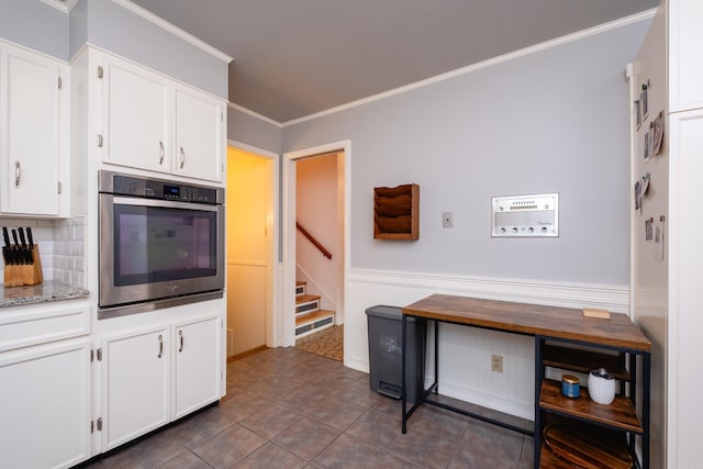 kitchen with stainless steel oven, backsplash, dark tile patterned flooring, ornamental molding, and white cabinetry