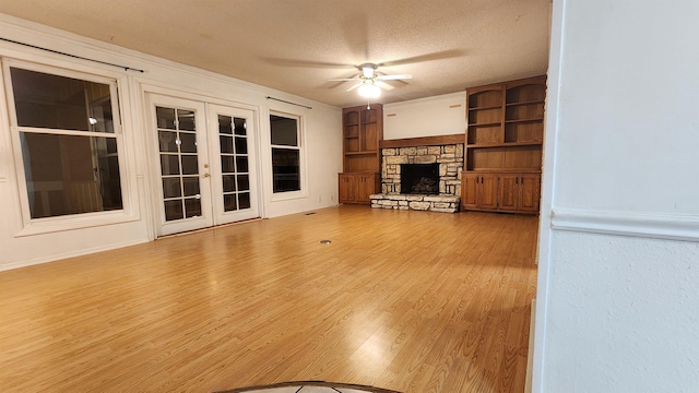 unfurnished living room with built in shelves, ceiling fan, french doors, a stone fireplace, and a textured ceiling