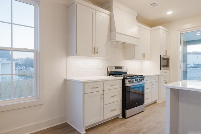 kitchen with light hardwood / wood-style flooring, decorative backsplash, white cabinets, custom range hood, and appliances with stainless steel finishes