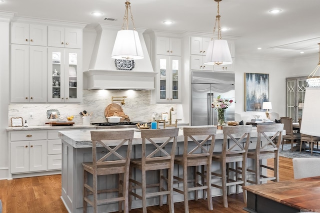 kitchen with a kitchen bar, custom range hood, a center island with sink, white cabinets, and built in fridge