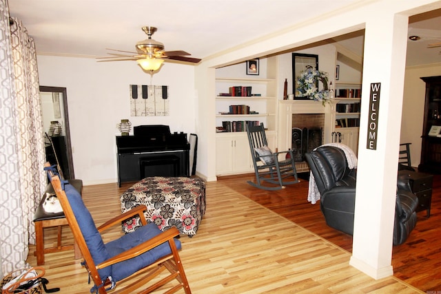 living room featuring a brick fireplace, built in shelves, ceiling fan, ornamental molding, and light hardwood / wood-style floors