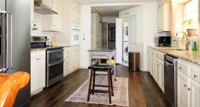 kitchen featuring sink, stainless steel appliances, stacked washing maching and dryer, light stone counters, and ventilation hood