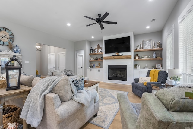 living room featuring built in shelves, light wood-type flooring, a large fireplace, and ceiling fan