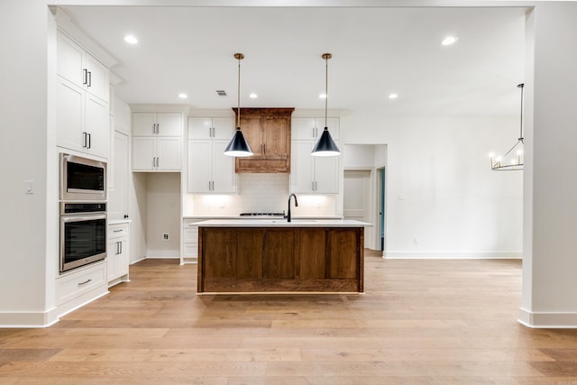 kitchen featuring stainless steel microwave, hanging light fixtures, a center island with sink, white cabinets, and light wood-type flooring