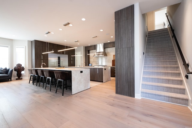 kitchen with light hardwood / wood-style flooring, an island with sink, wall chimney range hood, backsplash, and a kitchen breakfast bar