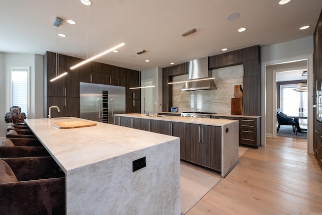 kitchen with sink, built in fridge, wall chimney exhaust hood, light hardwood / wood-style flooring, and an island with sink