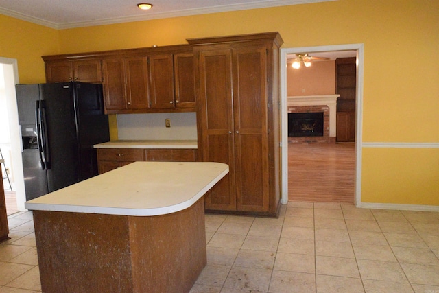 kitchen featuring black refrigerator with ice dispenser, a brick fireplace, a kitchen island, crown molding, and ceiling fan