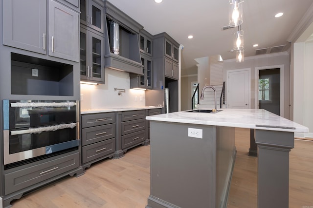 kitchen with sink, gray cabinets, ornamental molding, an island with sink, and decorative light fixtures