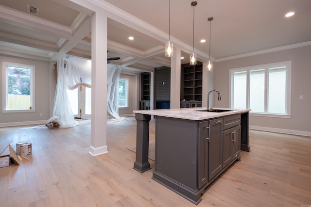 kitchen with coffered ceiling, sink, light stone counters, decorative light fixtures, and a kitchen island with sink