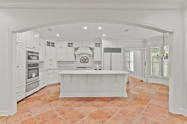 kitchen featuring white cabinets, backsplash, premium range hood, and an island with sink