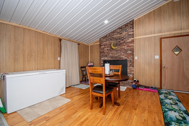 dining space with lofted ceiling, wooden walls, a brick fireplace, and light hardwood / wood-style flooring