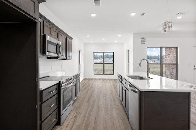 kitchen featuring sink, light wood-type flooring, an island with sink, hanging light fixtures, and appliances with stainless steel finishes