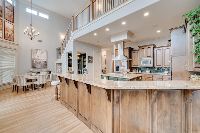 kitchen with wall chimney exhaust hood, kitchen peninsula, light stone counters, a breakfast bar, and stainless steel microwave