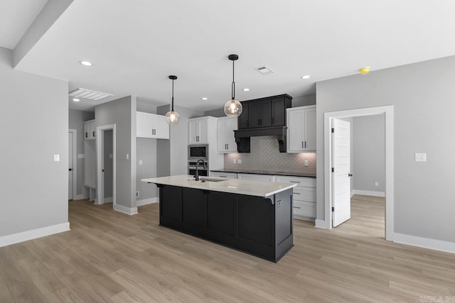 kitchen featuring an island with sink, stainless steel microwave, backsplash, light stone counters, and sink