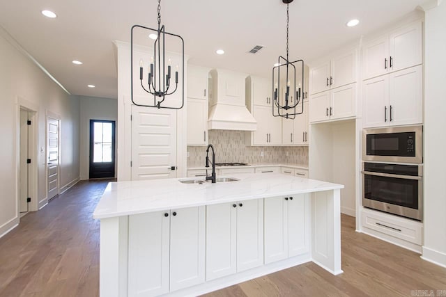 kitchen featuring a center island with sink, stainless steel oven, black microwave, custom range hood, and light stone counters