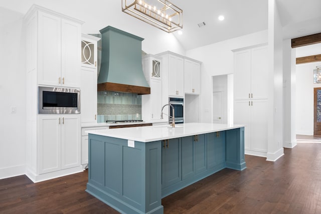 kitchen with appliances with stainless steel finishes, hanging light fixtures, white cabinets, a center island with sink, and custom exhaust hood