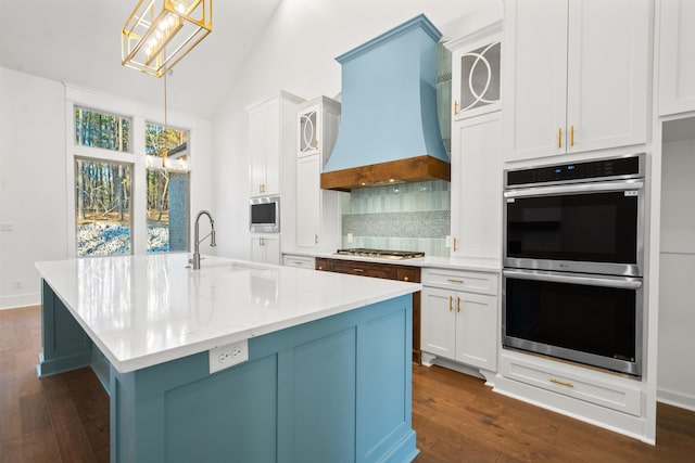 kitchen featuring pendant lighting, white cabinets, stainless steel appliances, custom range hood, and a center island with sink