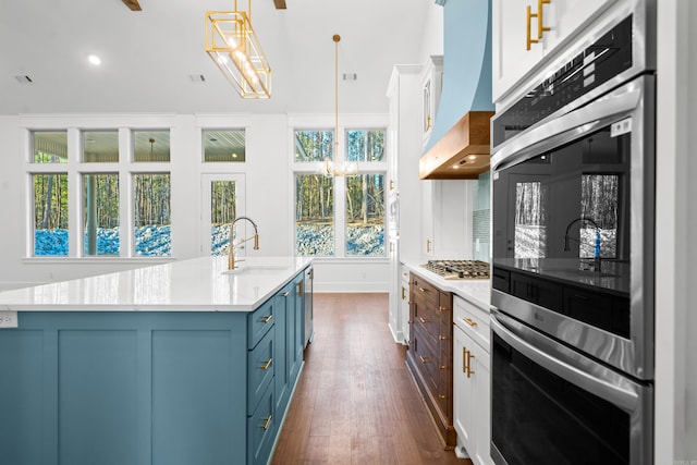 kitchen featuring appliances with stainless steel finishes, decorative light fixtures, white cabinetry, a kitchen island with sink, and custom range hood