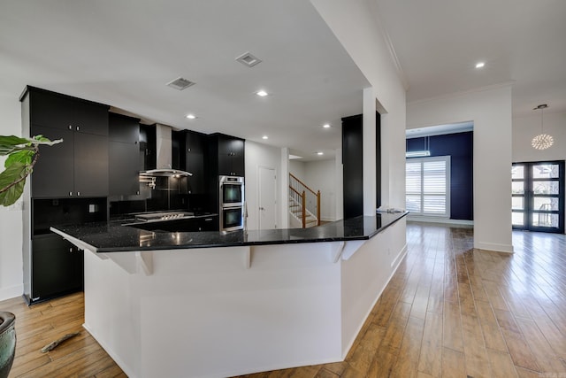 kitchen with pendant lighting, stainless steel double oven, wall chimney range hood, light hardwood / wood-style floors, and a kitchen breakfast bar