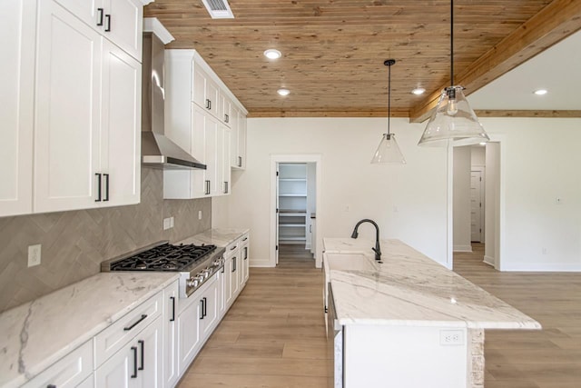 kitchen featuring decorative light fixtures, wall chimney range hood, an island with sink, light stone counters, and wooden ceiling