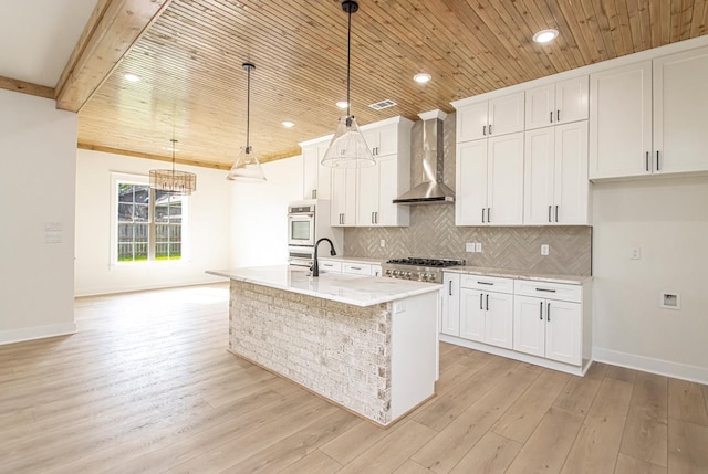 kitchen with white cabinetry, wall chimney range hood, an island with sink, hanging light fixtures, and light stone counters