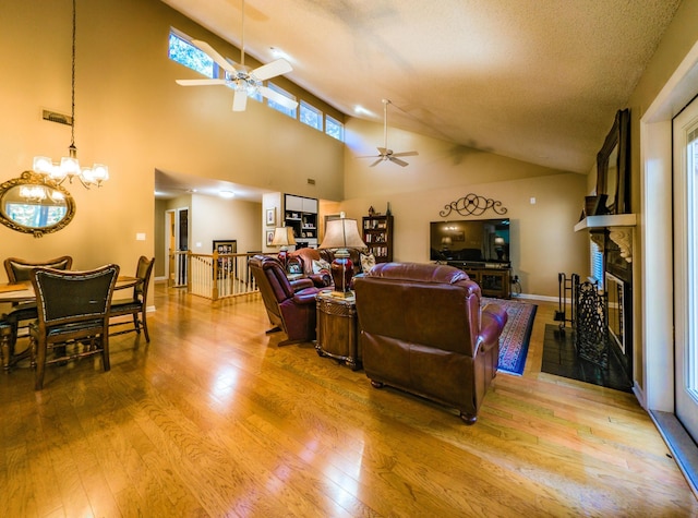 living room featuring ceiling fan with notable chandelier, a high ceiling, a textured ceiling, a stone fireplace, and light hardwood / wood-style floors