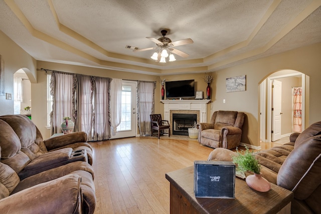 living room with a textured ceiling, hardwood / wood-style flooring, ceiling fan, a tray ceiling, and a tiled fireplace