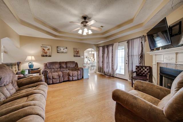 living room featuring a textured ceiling, a fireplace, a raised ceiling, light wood-type flooring, and ceiling fan