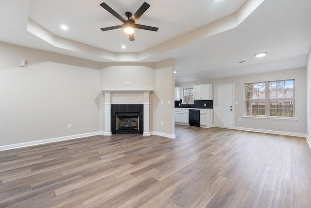 unfurnished living room with a raised ceiling, ceiling fan, a tile fireplace, and light hardwood / wood-style floors