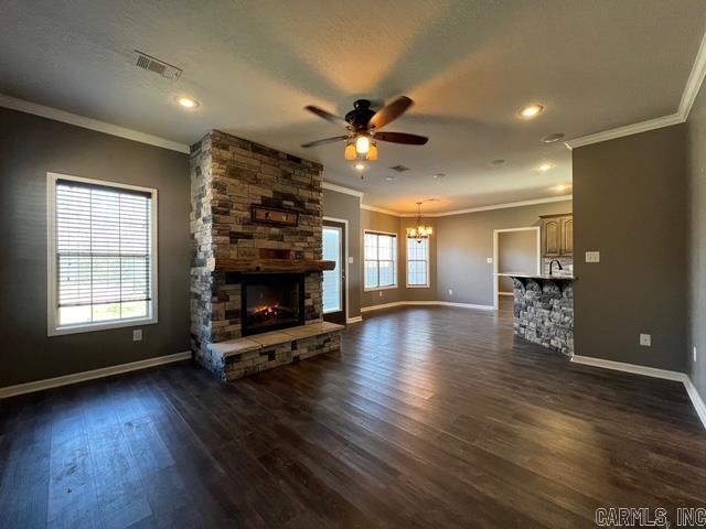 unfurnished living room featuring ceiling fan with notable chandelier, dark wood-type flooring, ornamental molding, and a stone fireplace