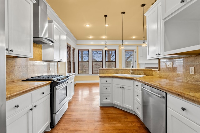 kitchen featuring crown molding, appliances with stainless steel finishes, white cabinetry, wall chimney range hood, and decorative light fixtures