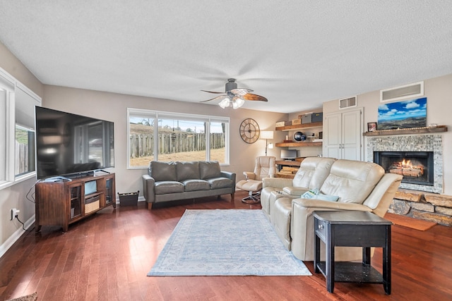 living room with ceiling fan, dark wood-type flooring, a textured ceiling, and a stone fireplace