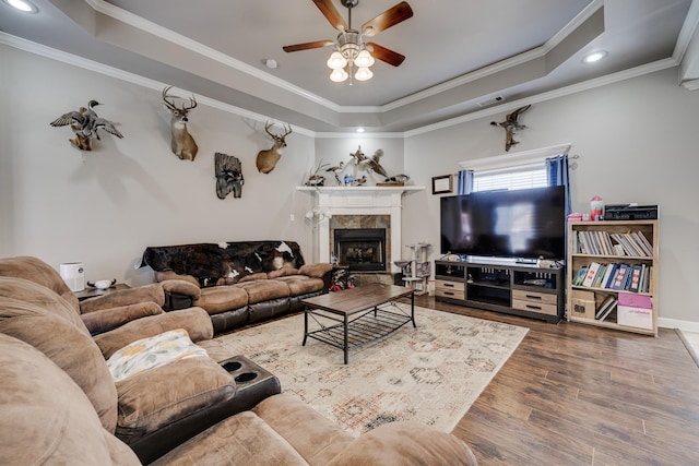 living room featuring ceiling fan, a raised ceiling, hardwood / wood-style flooring, a tile fireplace, and crown molding