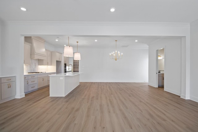 kitchen with hanging light fixtures, custom exhaust hood, sink, light wood-type flooring, and an island with sink