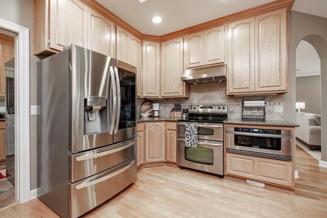 kitchen featuring light brown cabinetry, tasteful backsplash, dark stone countertops, stainless steel appliances, and light wood-type flooring