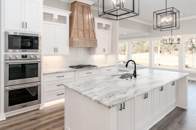 kitchen featuring premium range hood, an island with sink, sink, white cabinets, and hanging light fixtures