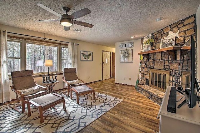 sitting room featuring ceiling fan, wood-type flooring, a stone fireplace, and a textured ceiling