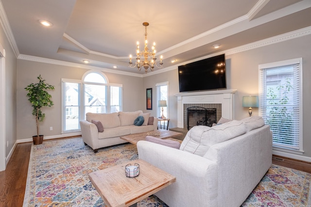 living room featuring wood-type flooring, a chandelier, ornamental molding, a raised ceiling, and a brick fireplace