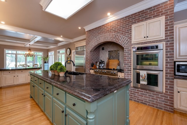 kitchen featuring an island with sink, sink, built in appliances, green cabinetry, and beam ceiling