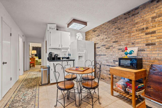 kitchen with brick wall, white cabinets, white refrigerator with ice dispenser, light tile patterned floors, and a textured ceiling