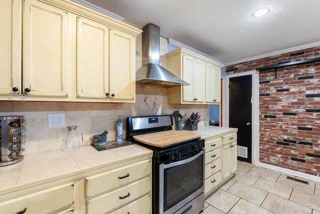 kitchen with brick wall, light tile patterned floors, wall chimney exhaust hood, stainless steel range oven, and cream cabinetry