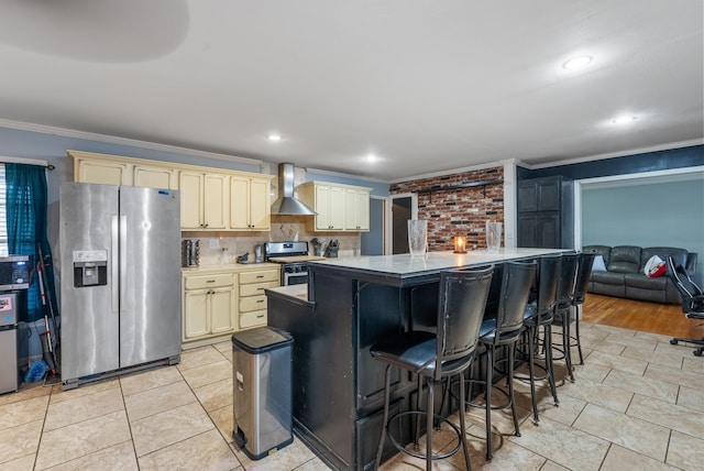 kitchen with crown molding, appliances with stainless steel finishes, cream cabinets, decorative backsplash, and wall chimney range hood