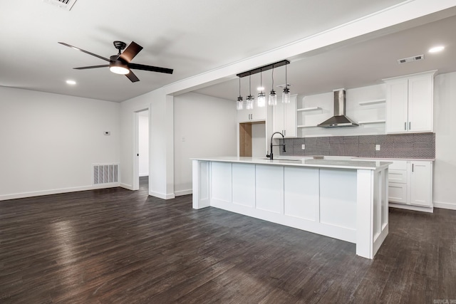 kitchen with wall chimney range hood, backsplash, white cabinets, and a center island with sink