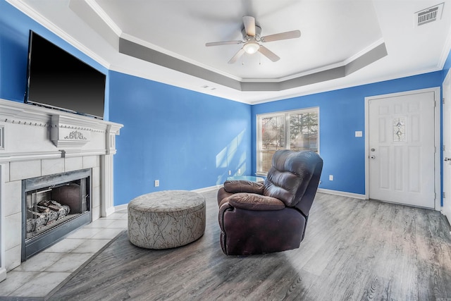 living room with a tile fireplace, ceiling fan, ornamental molding, a raised ceiling, and light wood-type flooring