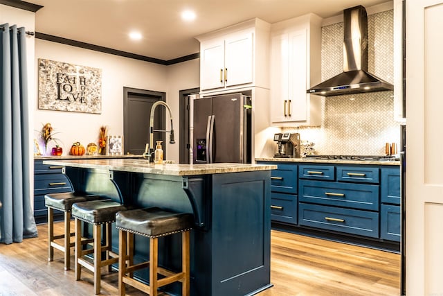 kitchen with wall chimney range hood, white cabinetry, stainless steel appliances, blue cabinets, and a center island with sink