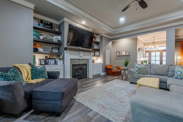 living room with a stone fireplace, ornamental molding, a tray ceiling, hardwood / wood-style floors, and ceiling fan with notable chandelier