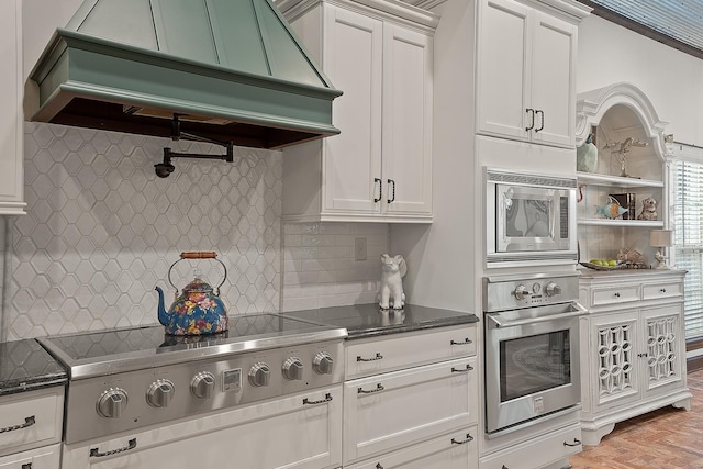 kitchen with white cabinetry, stainless steel appliances, tasteful backsplash, custom exhaust hood, and dark stone counters